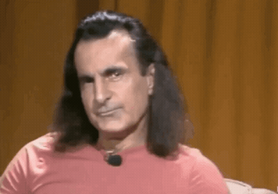A Man With Long Hair And A Pink Shirt Is Sitting In Front Of A Brown Curtain . GIF