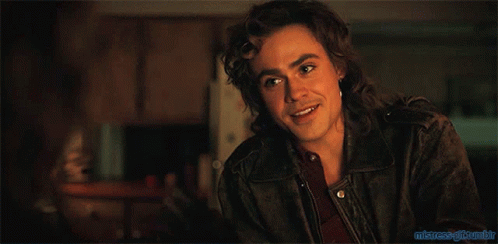 A Man With Long Hair And A Ring On His Finger Is Sitting At A Table In A Dark Room GIF