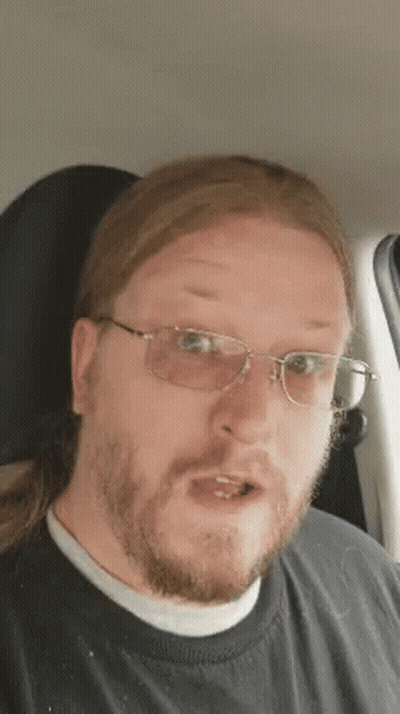 A Man With Long Hair And Glasses Is Sitting In A Car And Making A Funny Face . GIF