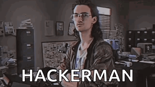 A Man With Long Hair And Glasses Is Standing In A Room With The Word Hackerman Written On The Screen . GIF