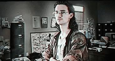 A Man With Long Hair And Glasses Is Standing In Front Of A Desk In An Office . GIF