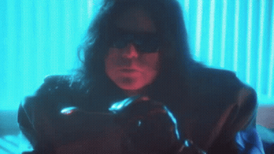 A Man With Long Hair And Sunglasses Is Sitting In Front Of A Blue Wall GIF