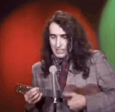 A Man With Long Hair Is Holding A Guitar And Speaking Into A Microphone . GIF