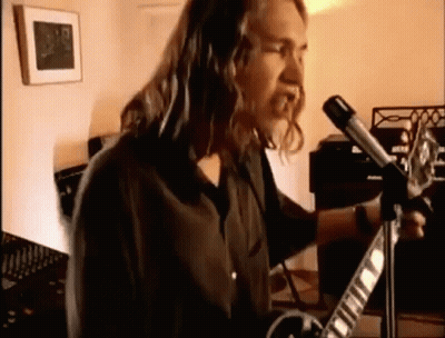 A Man With Long Hair Is Playing A Guitar In Front Of A Microphone In A Room . GIF