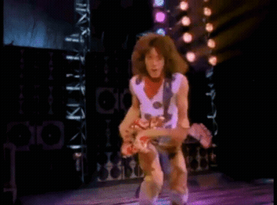 A Man With Long Hair Is Playing A Guitar On A Stage With Lights Behind Him . GIF