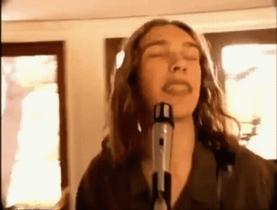 A Man With Long Hair Is Singing Into A Microphone In A Room . GIF