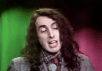 A Man With Long Hair Is Singing Into A Microphone In Front Of A Green And Pink Background . GIF