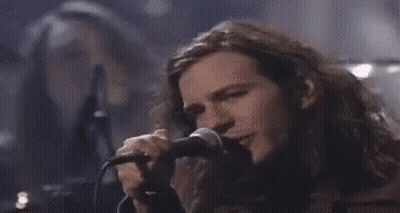 A Man With Long Hair Is Singing Into A Microphone While Another Man Looks On . GIF