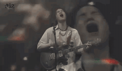 A Man With Long Hair Is Singing Into A Microphone While Making A Funny Face . GIF