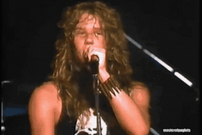 A Man With Long Hair Is Singing Into A Microphone While Standing On A Stage . GIF