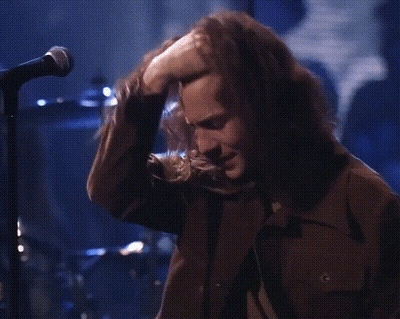 A Man With Long Hair Is Singing Into A Microphone While Touching His Nose . GIF