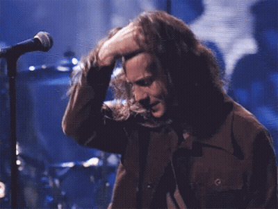 A Man With Long Hair Is Singing Into A Microphone While Wearing A Brown Jacket GIF