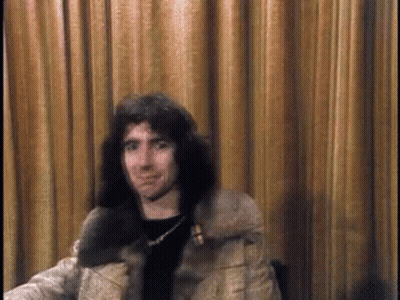 A Man With Long Hair Is Sitting In Front Of A Brown Curtain GIF