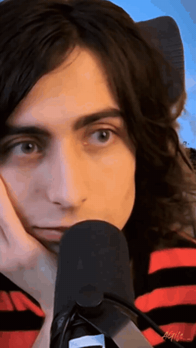 A Man With Long Hair Is Sitting In Front Of A Microphone With His Hand On His Chin . GIF