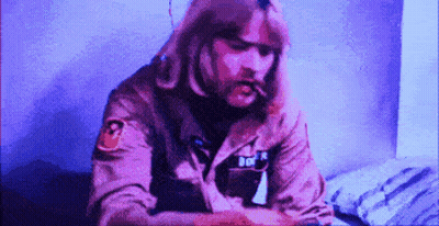 A Man With Long Hair Is Sitting On A Bed Smoking A Cigarette GIF