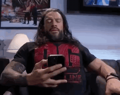 A Man With Long Hair Is Sitting On A Couch Looking At His Phone . GIF