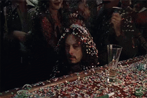 A Man With Long Hair Is Sitting On A Table Covered In Confetti And A Glass . GIF