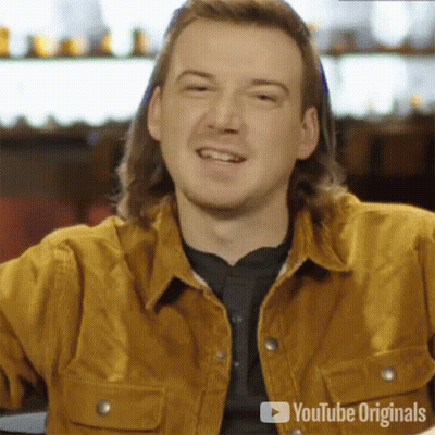 A Man With Long Hair Is Smiling And Wearing A Brown Jacket . GIF
