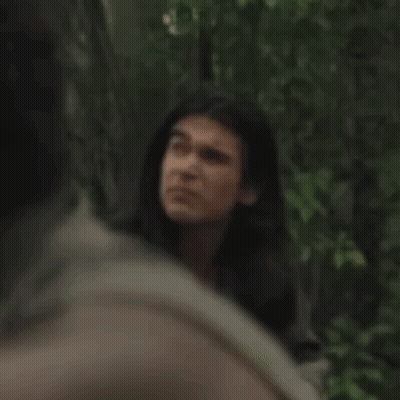 A Man With Long Hair Is Standing In A Forest Looking At Another Man . GIF