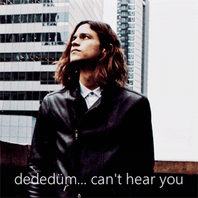A Man With Long Hair Is Standing In Front Of A Building With The Words Dededum Can 'T Hear You Written Below Him GIF