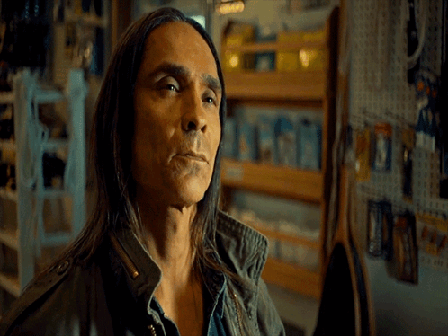 A Man With Long Hair Is Standing In Front Of A Wall That Says Okay GIF