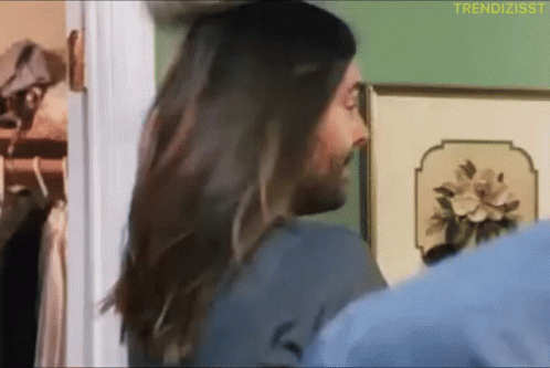 A Man With Long Hair Is Standing In Front Of A Wall With A Picture Of Flowers On It GIF