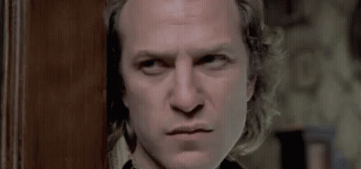 A Man With Long Hair Is Standing In Front Of A Wooden Door Looking At The Camera . GIF