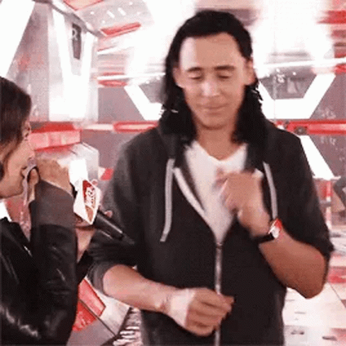 A Man With Long Hair Is Standing Next To A Woman Holding A Microphone And Talking To Her . GIF