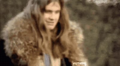 A Man With Long Hair Is Wearing A Fur Coat And Holding A Glass Of Water . GIF