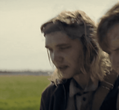 A Man With Long Hair Wearing Headphones Stands Next To A Woman In A Field GIF