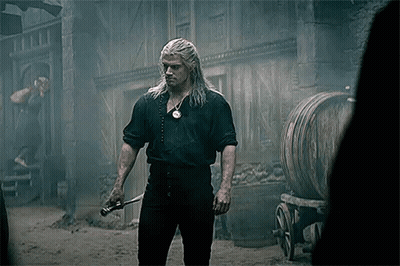 A Man With Long White Hair Is Holding A Sword In Front Of A Building And Barrels . GIF