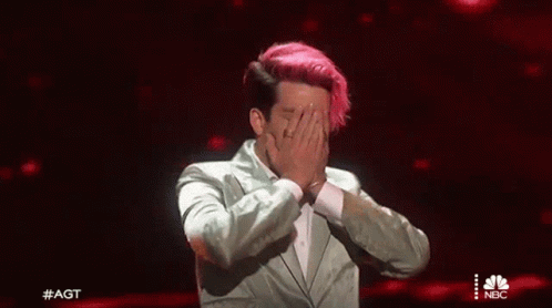 A Man With Pink Hair Is Covering His Face With His Hands While Wearing A Suit . GIF