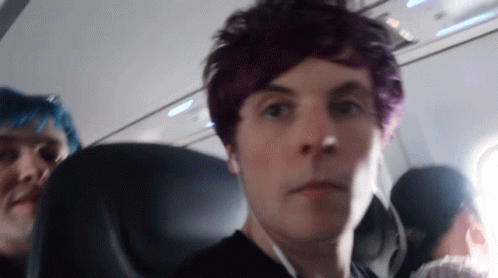 A Man With Purple Hair Looks At The Camera While Sitting On An Airplane GIF