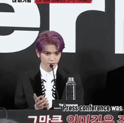 A Man With Purple Hair Speaking Into A Microphone In Front Of A Sign That Says Press Conference GIF