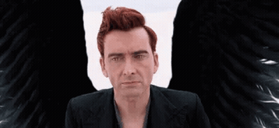 A Man With Red Hair And Black Wings Is Sitting In Front Of A White Wall . GIF