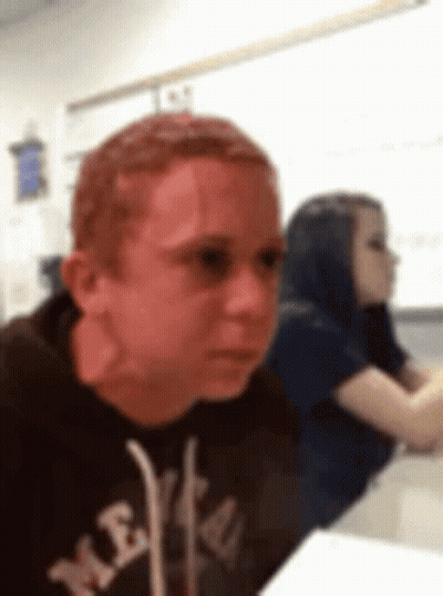 A Man With Red Hair Is Sitting At A Desk In A Classroom With A Woman In The Background . GIF