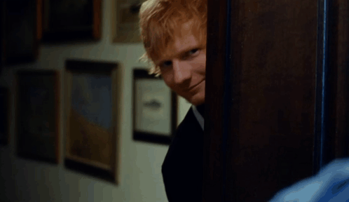 A Man With Red Hair Is Smiling While Peeking Out Of A Doorway GIF