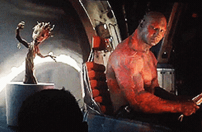A Man With Red Paint On His Body Is Sitting In A Chair Next To A Statue Of Groot . GIF