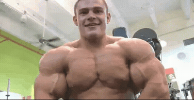 A Man With Very Large Muscles Is Standing In A Gym And Smiling At The Camera . GIF