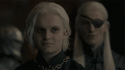 A Man With White Hair And A Necklace Is Standing Next To Another Man With A Blindfold On His Eye GIF