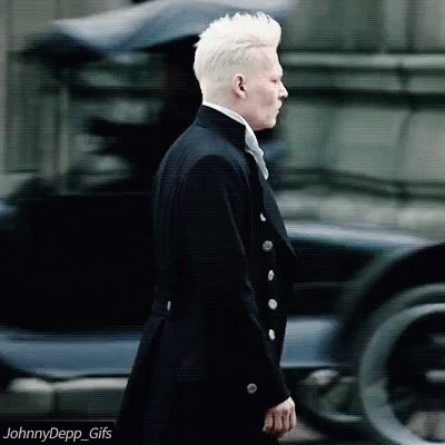 A Man With White Hair Is Wearing A Black Suit And Tie And Walking Down The Street . GIF