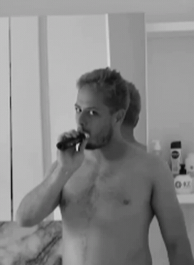 A Man Without A Shirt Is Brushing His Teeth In Front Of A Mirror . GIF