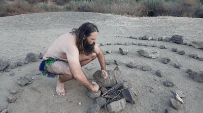 A Man Without A Shirt Is Kneeling In The Dirt Near A Rock Labyrinth GIF