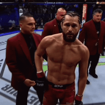 A Man Without A Shirt Is Standing In A Boxing Ring Surrounded By Men In Red Suits . GIF