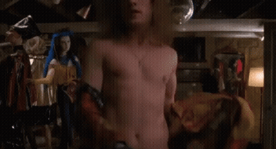 A Man Without A Shirt Is Standing In A Room With A Woman In A Costume Behind Him . GIF