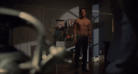 A Man Without A Shirt Is Standing In A Room With Posters On The Wall GIF