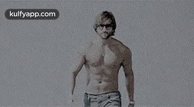 A Man Without A Shirt Is Standing On A Beach Wearing Sunglasses . GIF