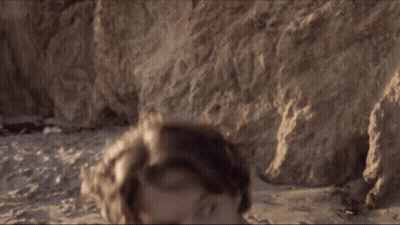 A Man Without A Shirt Is Standing On The Beach Looking At The Camera GIF