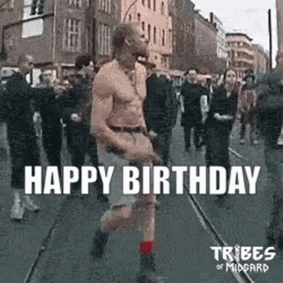 A Man Without A Shirt Is Walking Down A Street In Front Of A Crowd Of People . GIF