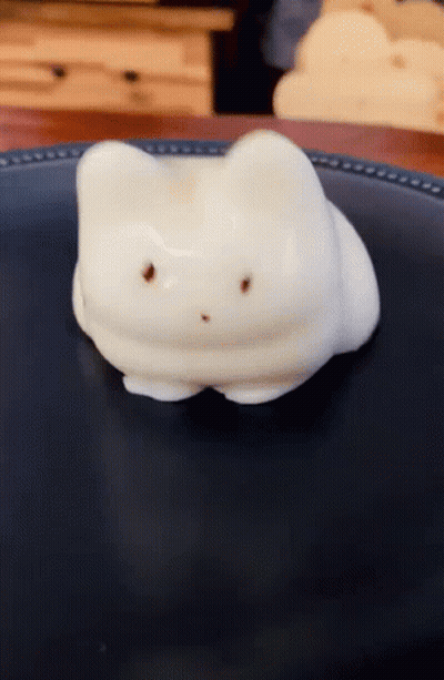 A Marshmallow In The Shape Of A Cat Laying On A Black Plate GIF